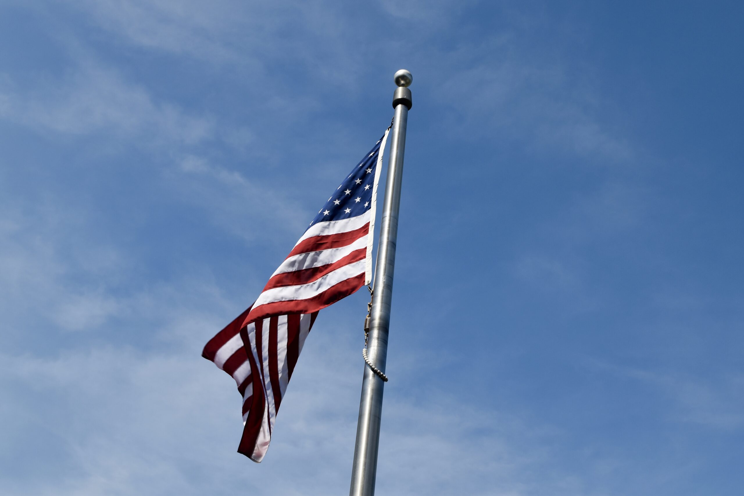 Low angle view of the American flag near trees under a blue cloudy sky and sunlight