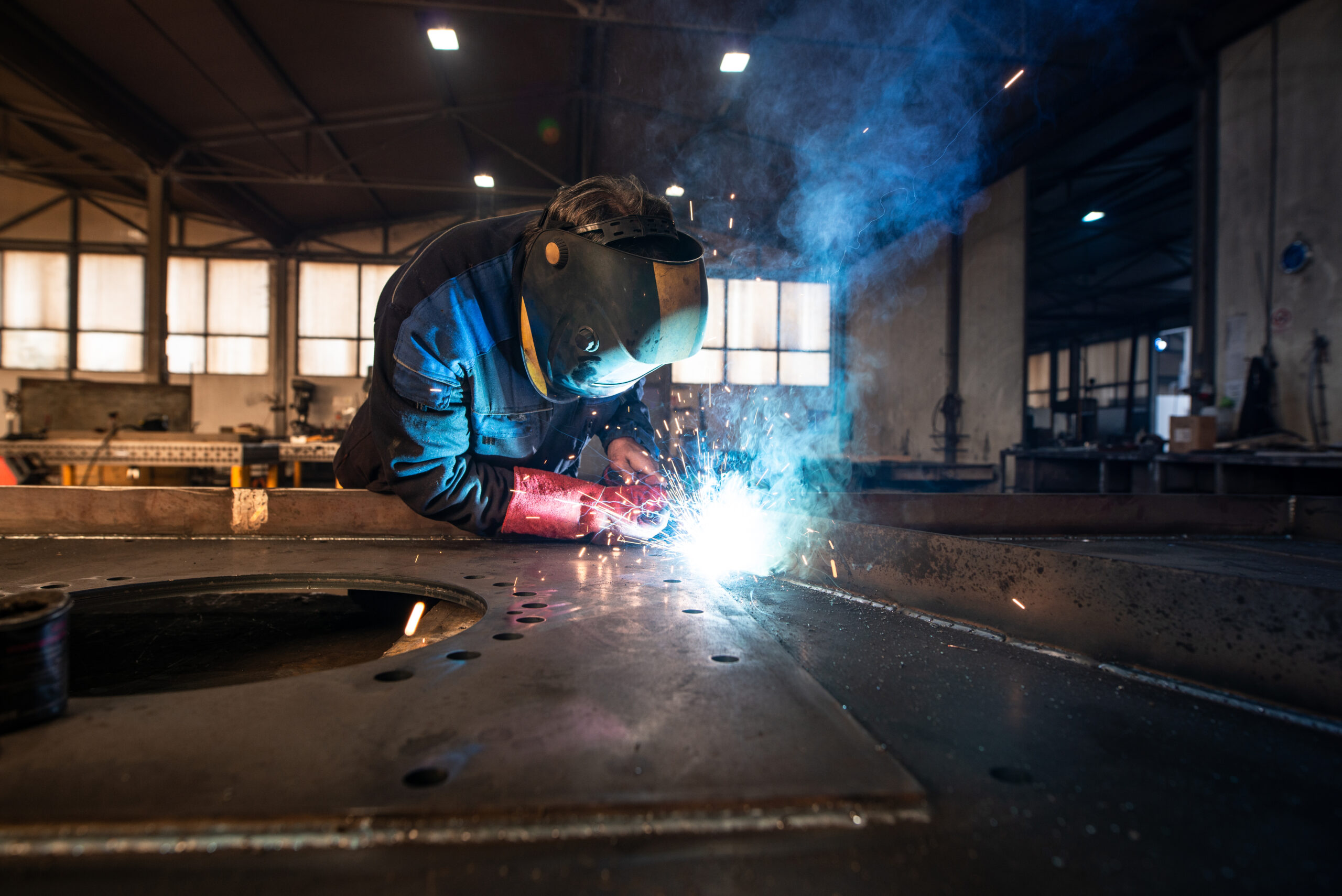 Professional industrial welder welding metal parts in metalworking factory. Wearing protective gear.